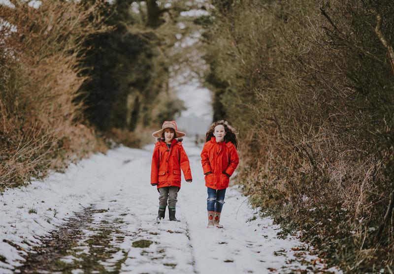 Two children walking on a snow-covered path in the winter, surrounded by bare trees and shrubbery. Both children are dressed warmly in bright red coats and are wearing boots suitable for the snow. One child has a hat on, and they both seem to be enjoying their walk through this chilly, serene landscape.