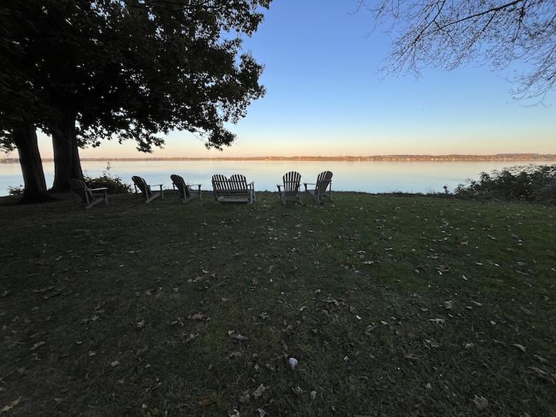 Six Adirondack chairs face a glassy lake at dusk, fallen leaves dotting the lawn beneath a massive oak, distant shore glowing warm