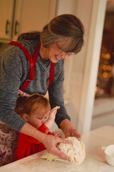 A grandmother in a red apron and a gray sweatshirt kneads bread dough, standing over her toddler-aged granddaughter, who is helping her enthusiastically