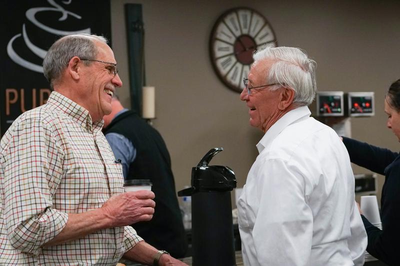 Two older gentlemen happily converse in front of a coffee station, their smiles reflecting the joy of friendship and good company.