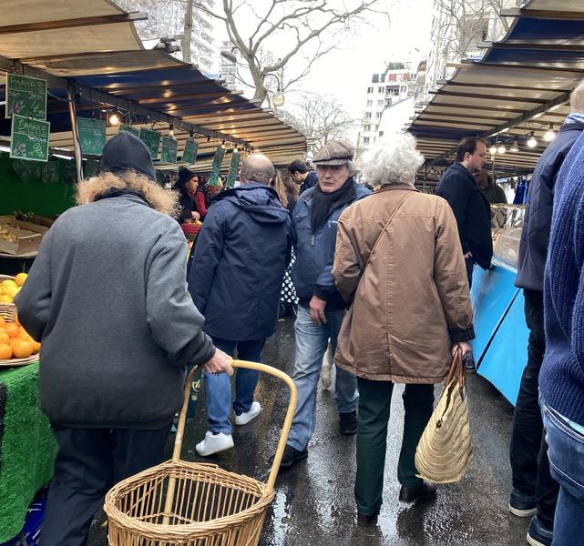 A bustling outdoor market scene in an urban environment with buildings visible in the background. People are walking and shopping. One person is carrying a wicker basket, while another is holding a straw bag. Several market stalls are visible on both sides, displaying fruits and other goods. The ground appears wet, suggesting recent rain.