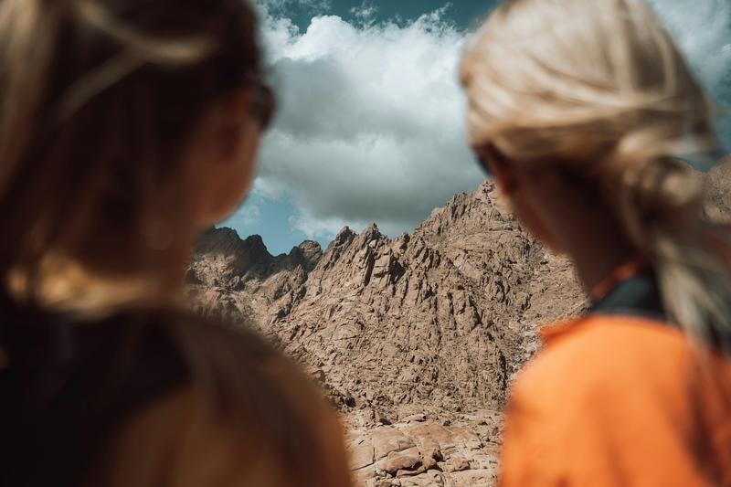 The image captures two individuals from behind, one with dark hair and the other with blonde hair in a braid, both gazing towards a distant, craggy mountain range under a partly cloudy sky. The focus is on the stark mountains, while the people appear blurred in the foreground.