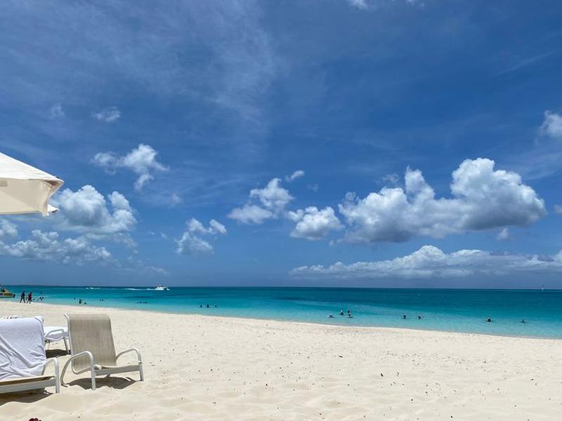 Two beach lounge chairs and a sun umbrella on white sand beside calm turquoise water under a blue sky with scattered clouds