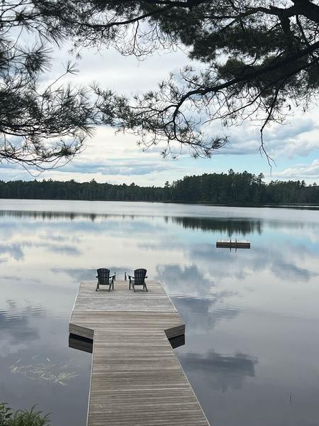 Two empty Adirondack chairs at the end of a wooden dock on a still lake, framed by pine branches and reflected clouds