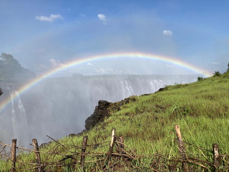 A vibrant rainbow arcing across the sky. Below, a vast waterfall cascades down a cliff, shrouded in mist. In the foreground, grassy African savannah receeds behind simple wooden fence.