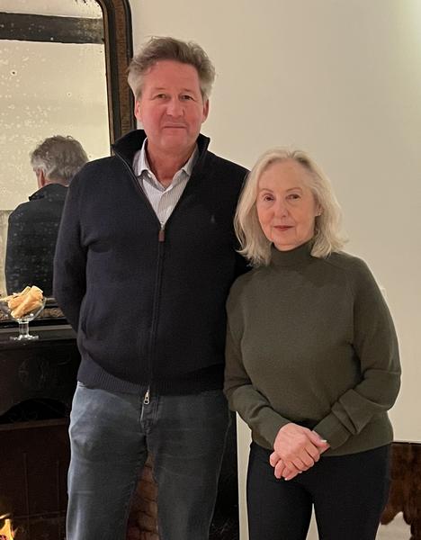 Prof. Dr. S.P.J. van Alphen, standing on the left, wearing a dark zip-up sweater over a checkered shirt, paired with jeans. Dr. Erlene Rosowsky, Psy.D., on the right, is wearing a dark olive green sweater and black pants. Both are looking directly at the camera with serious expressions. In the background, there's a mirror reflecting the back of the man, and a fireplace with a lit fire, adding warmth to the room.