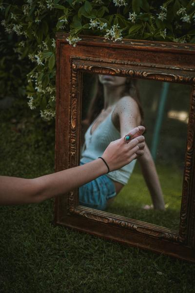 A woman touches her reflection in a mirror with an elaborate frame that leans against a shrub