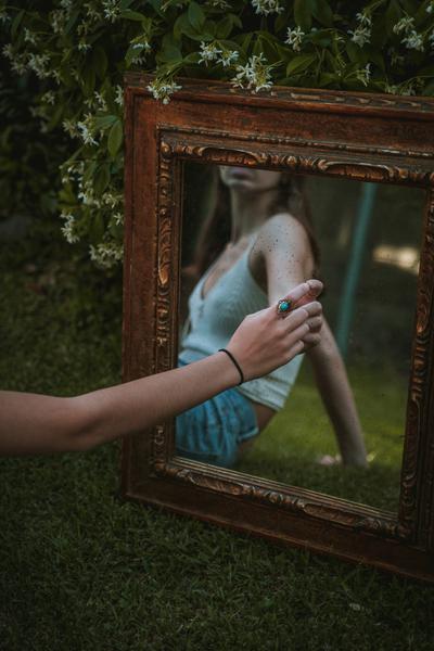 A woman touches her reflection in a mirror with an elaborate frame that leans against a shrub