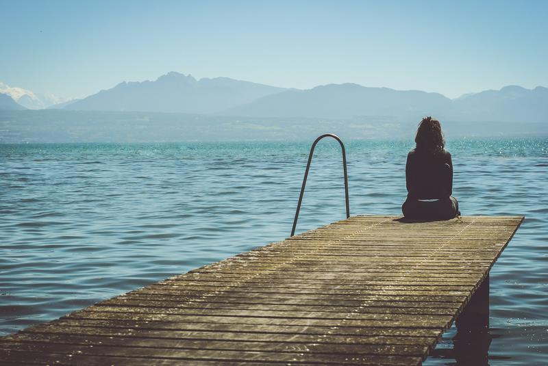 A lonely woman sits at the end of a dock overlooking a lake with fog-shrouded mountains in the background